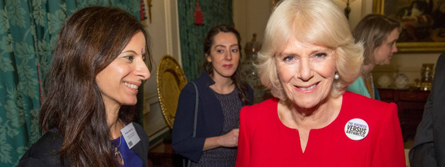 Smiling Camilla wearing a red dress and Arthritis UK badge standing beside a female researcher