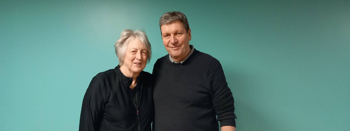 Smiling Paul wearing a navy jumper standing beside smiling Anne wearing a navy shirt.