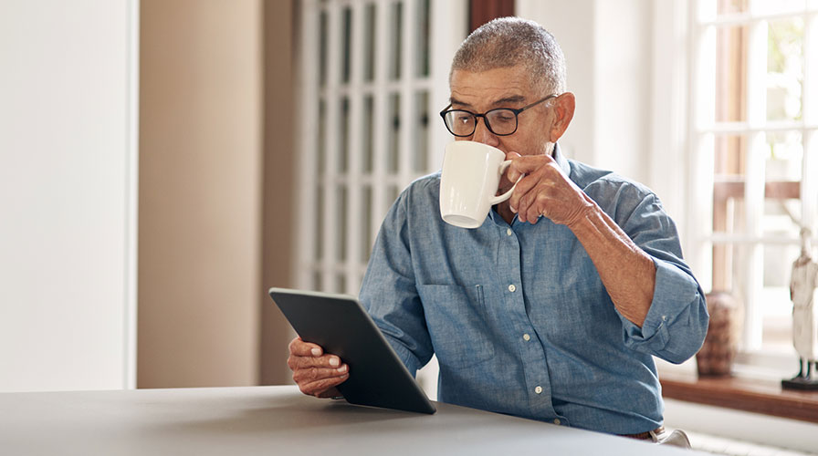 A man sitting in his kitchen drinking coffee while watching a live online session on his tablet.