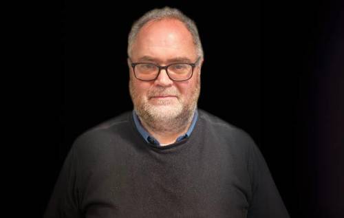Robin, who wears glasses and has a beard, waring a grey jumper in front of a black background